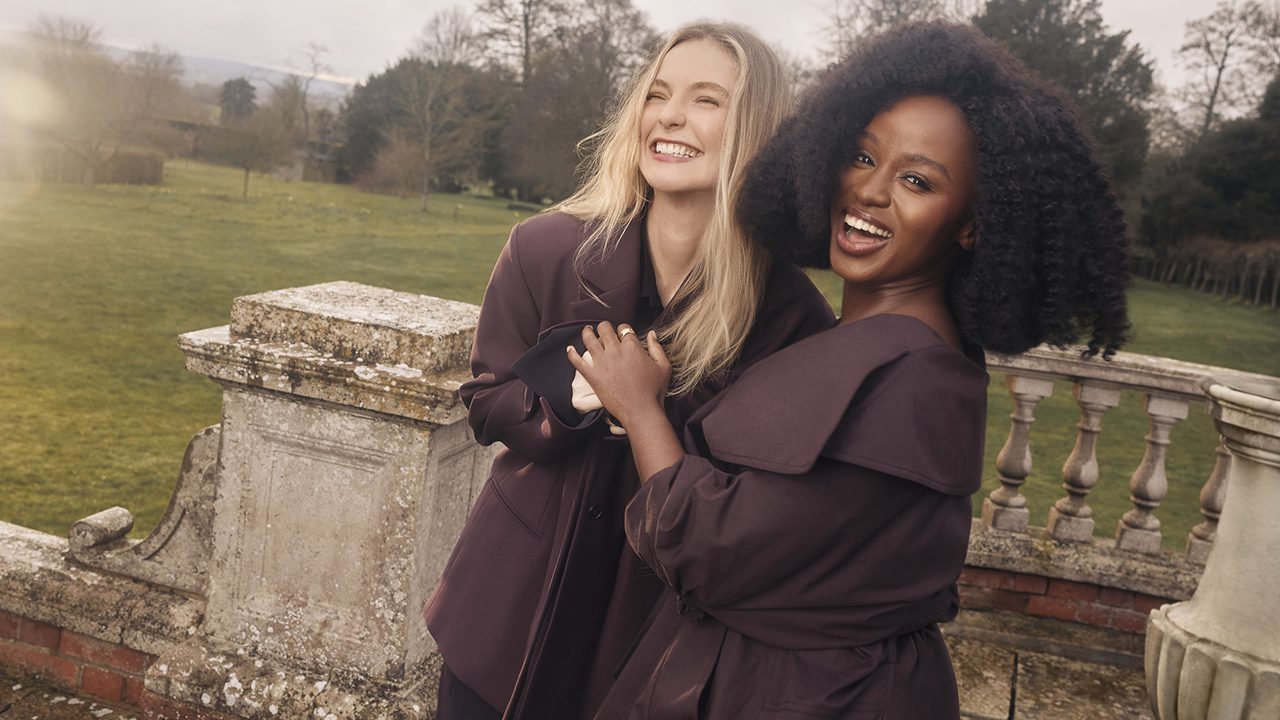 "Two women standing outdoors on grass near a stone railing, both wearing long dark coats, smiling and holding hands, with trees and cloudy sky in the background in a serene park or garden setting."
